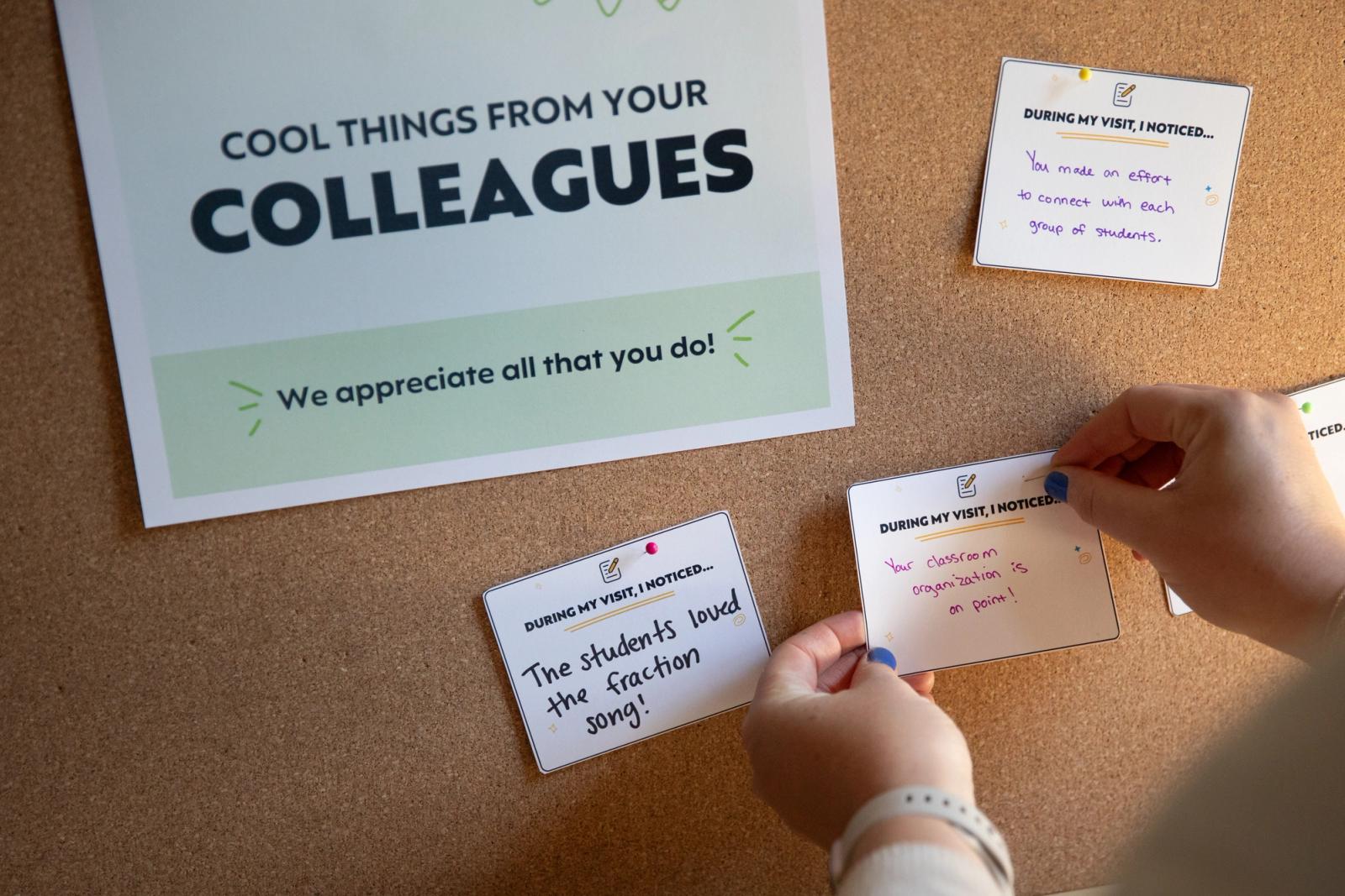 A bulletin board with several pieces of paper pinned to it. Someone reaches in to pin a notecard on the board.