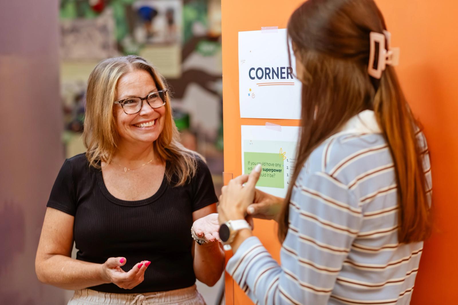 Two women stand in front of a wall with posters that say "corner 1" on it. 