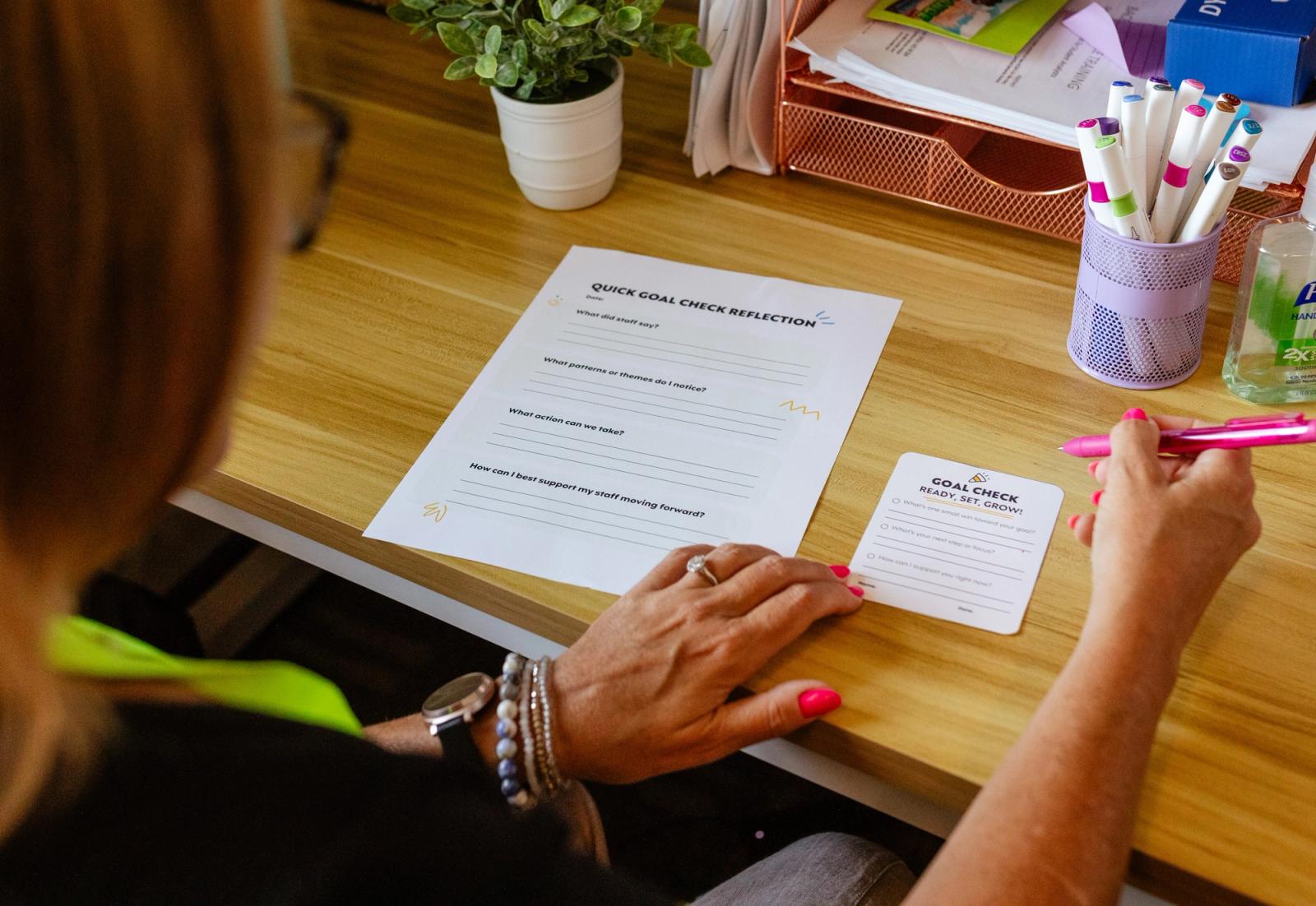 A woman sits at a wooden desk with a jar of markers and potted plant on it. She has a piece of paper and notecard in front of her and she is writing on them.
