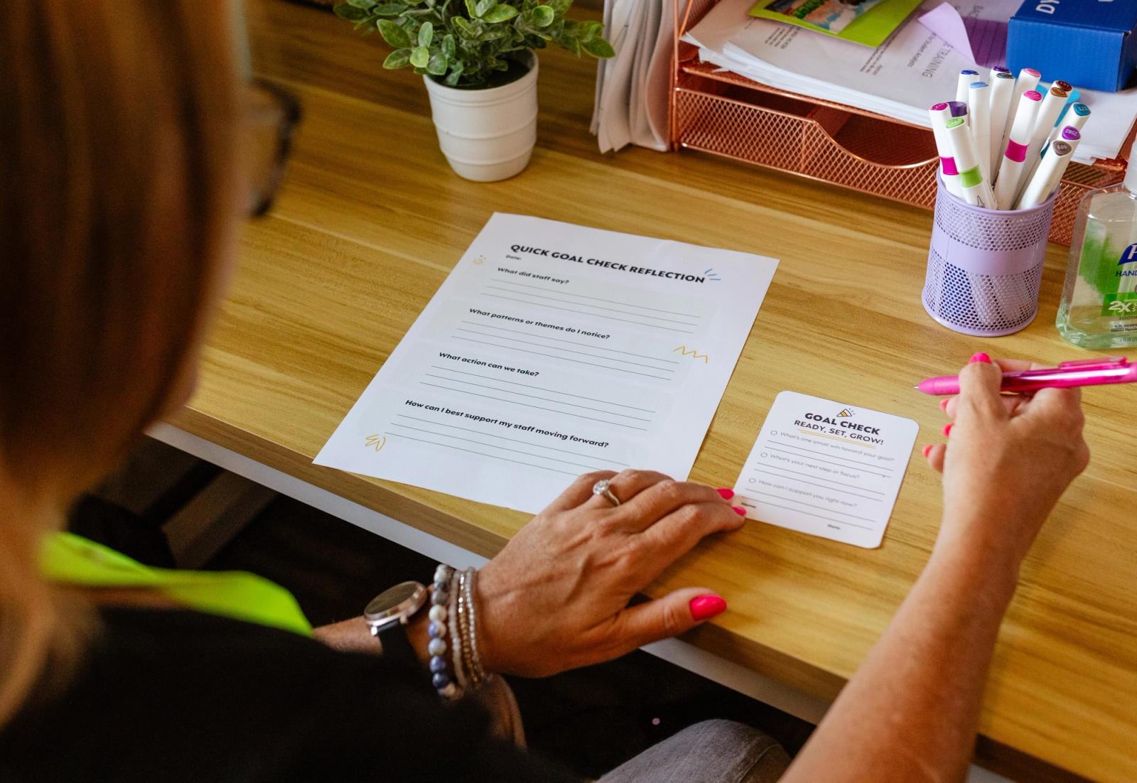 A woman sits at a wooden desk with a jar of markers and potted plant on it. She has a piece of paper and notecard in front of her and she is writing on them.