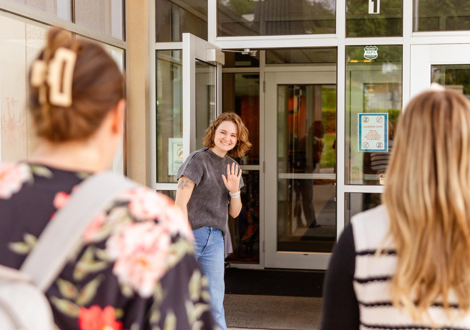 A woman stands in a doorway to a school waving. Two women walk toward her.