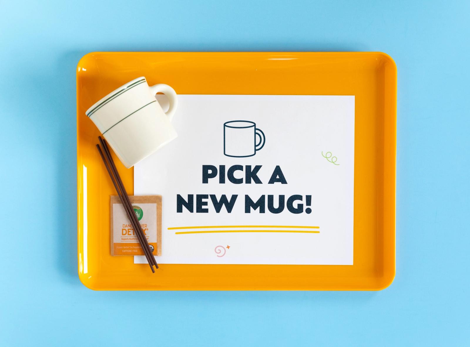 A yellow tray with a poster that says "pick a new mug", coffee cup, and tea bag sits on a light blue background.