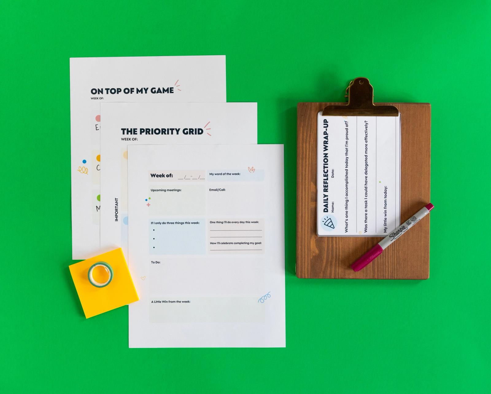 Three sheets of paper lay on a green background. Beside them are yellow sticky notes and a small clipboard with notecards and a pen on it.