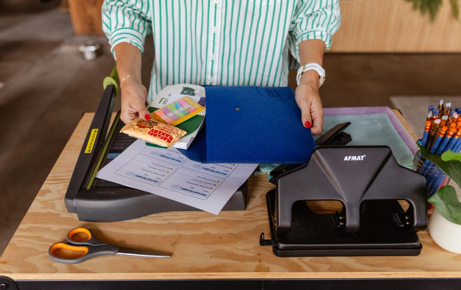 Someone stands in front of a wood table with paper cuttters and office supplies on it. They place items in a blue folder.