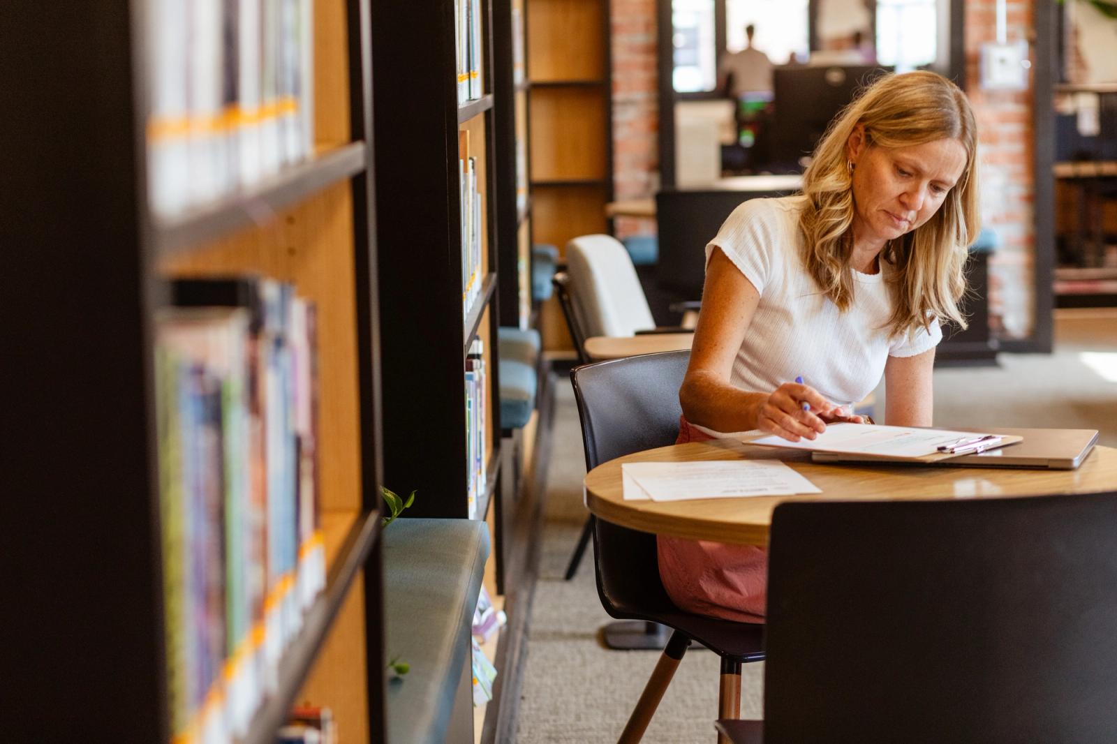 A woman sits next to bookshelves as a table writing on a piece of paper.