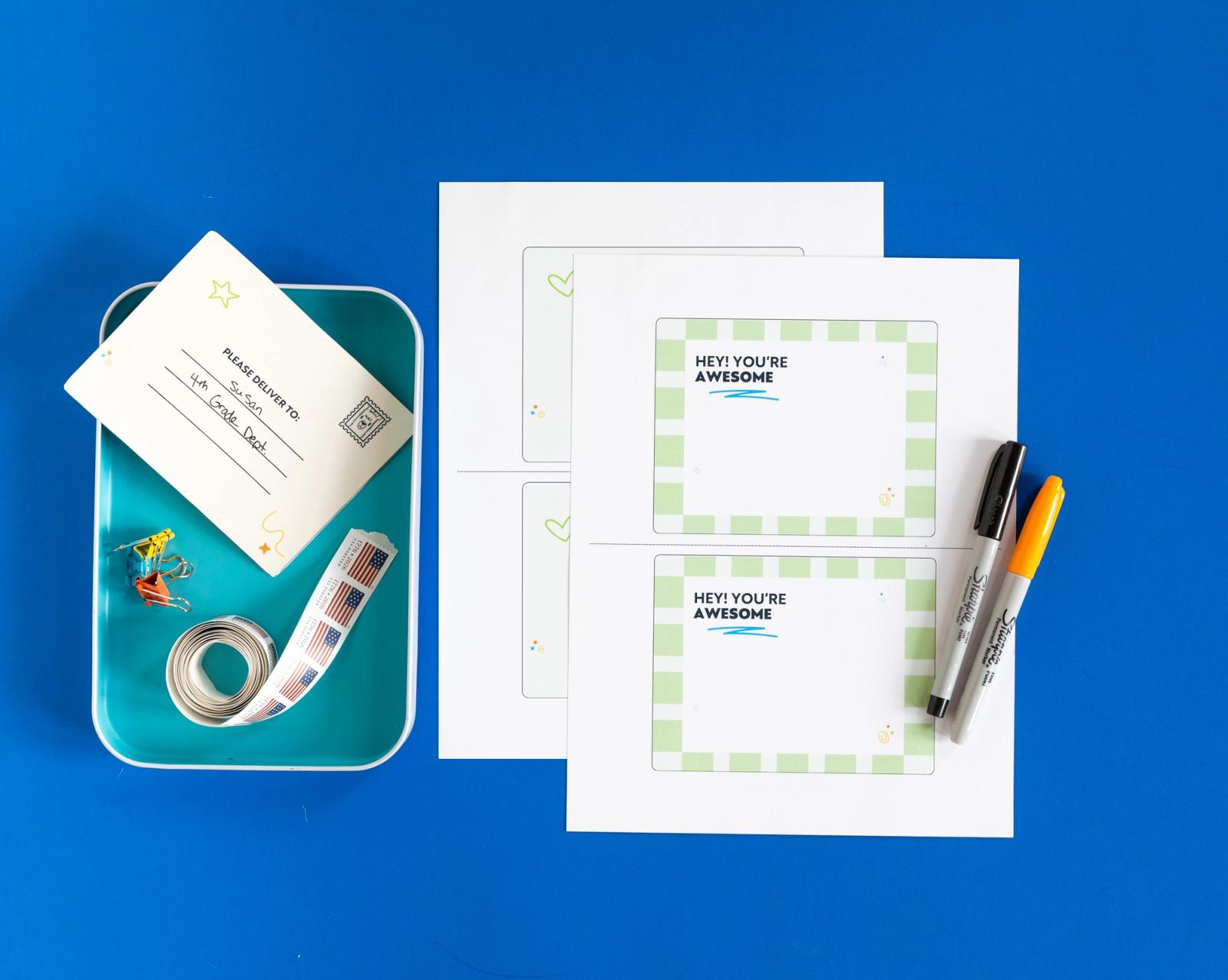Two sheets of paper and two markers lay on a blue background. Next to them is a blue container with a postcards and roll of stamps in it.