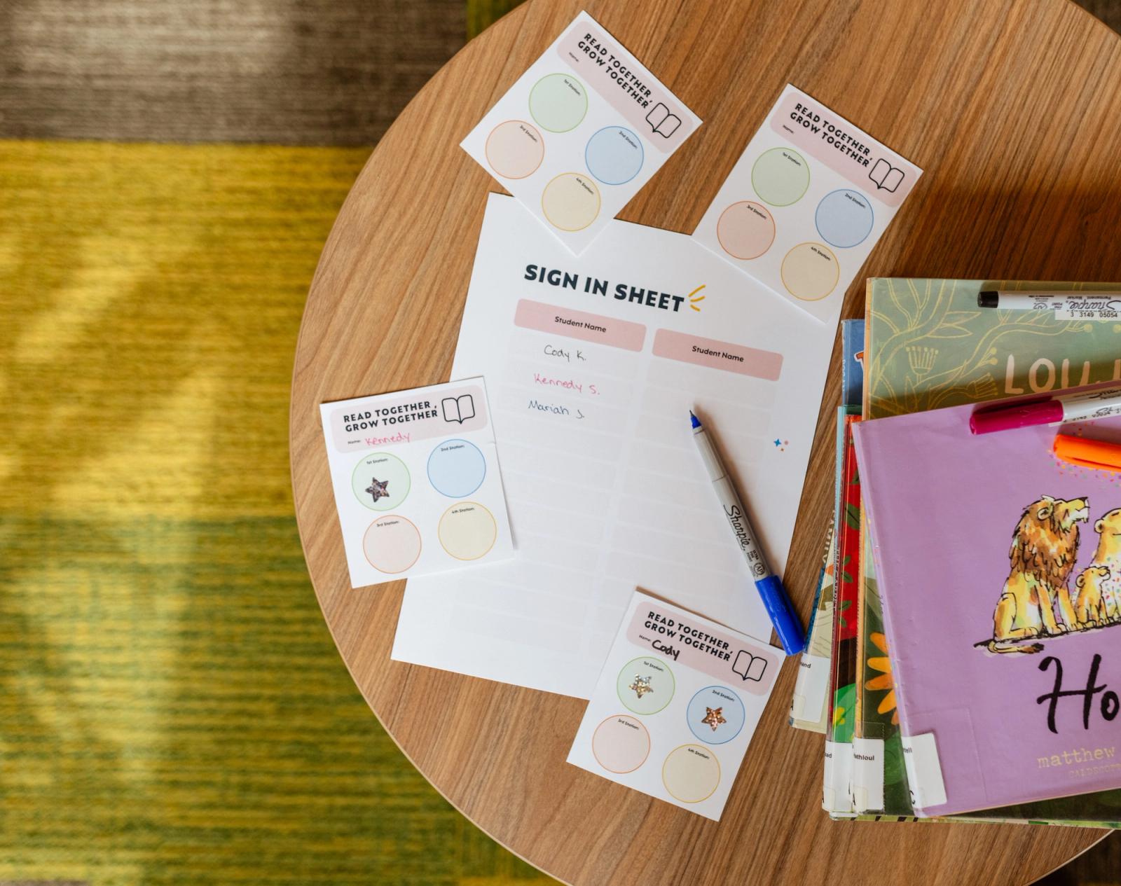 On a small round table lays a stack of children's books. Next to it is a sign-in sheet, and small pieces of paper with colorful papers on them. A colorful rug is in the background.