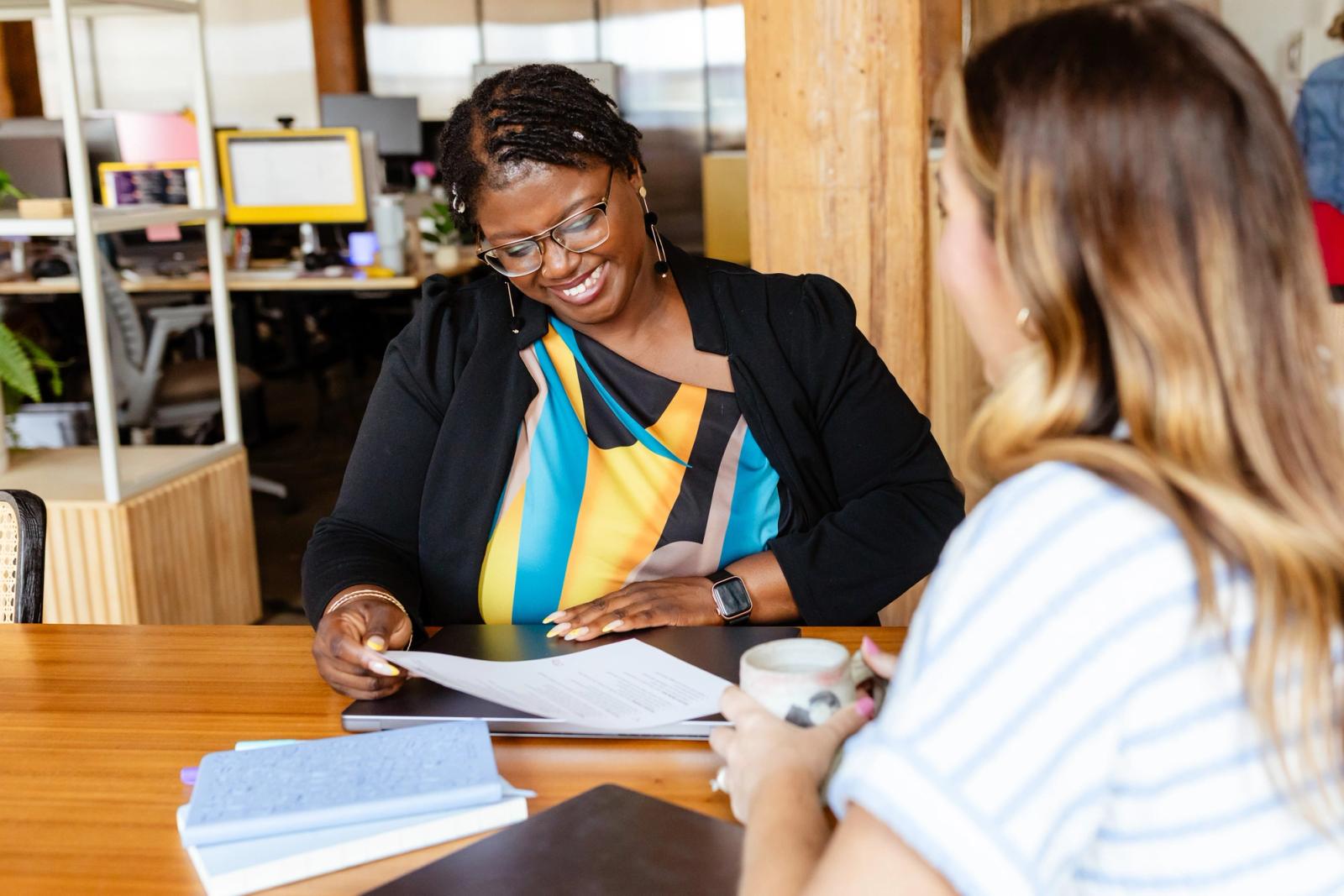 Two women sit across from each other at a table. Papers, laptops and notebooks lay on the table.