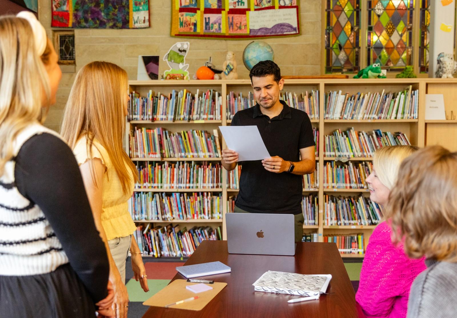 A man stands in front of a table in a library reading off a piece of paper. At the table, two women stand and two women sit.