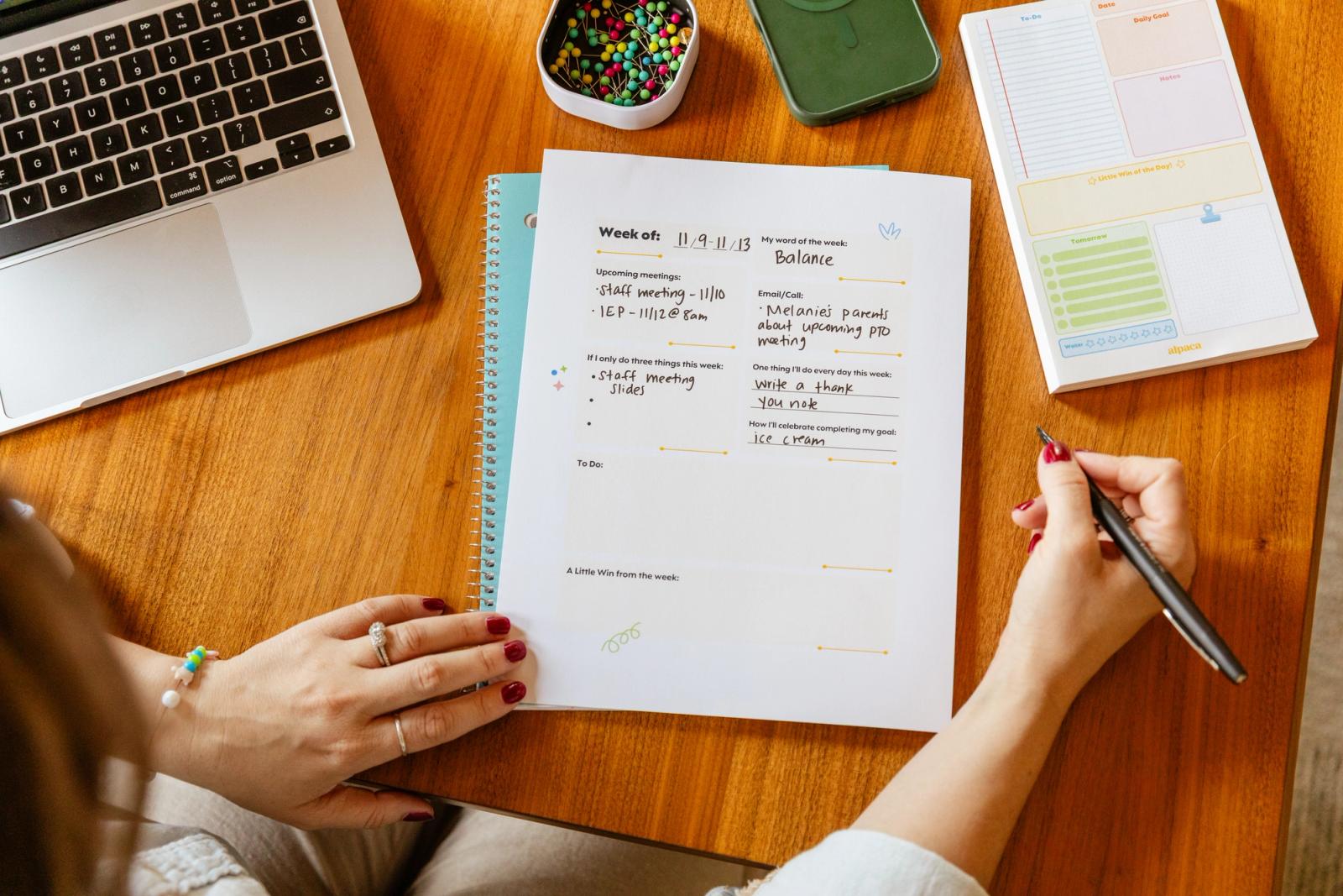 A hand writes on a piece of paper that lays on a wood table with an open laptop and notebook nearby.