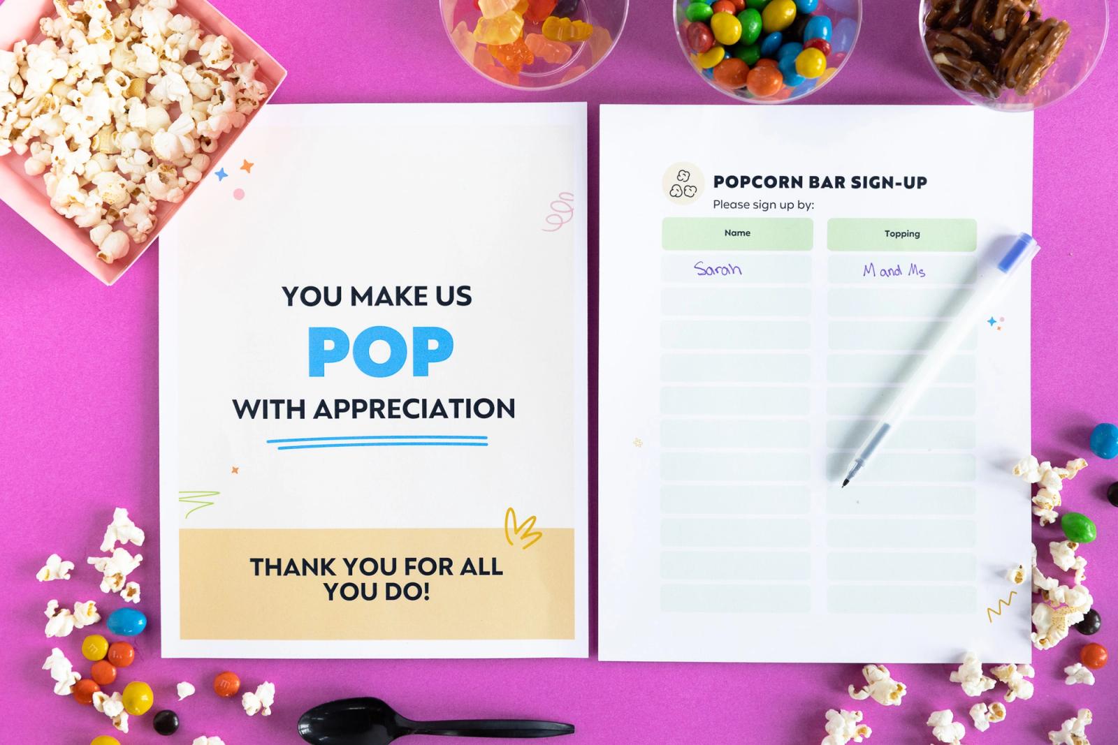 Two sheets of paper and a pen lay on a magenta background. Popcorn, candy, and toppings lay around them.