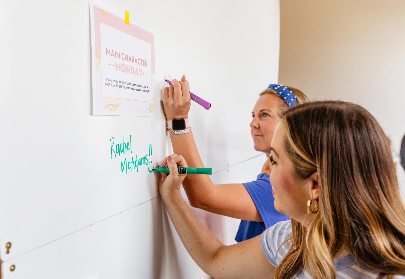Two women write on a whiteboard underneath a piece of paper taped to it.