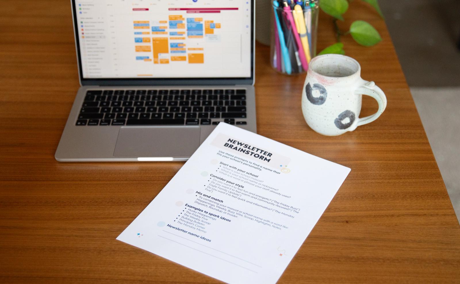 A computer with an open calendar on the screen sits on a table with a coffee cup and cup of pens on it. A paper with newsletter tips lays on top of the keyboard.