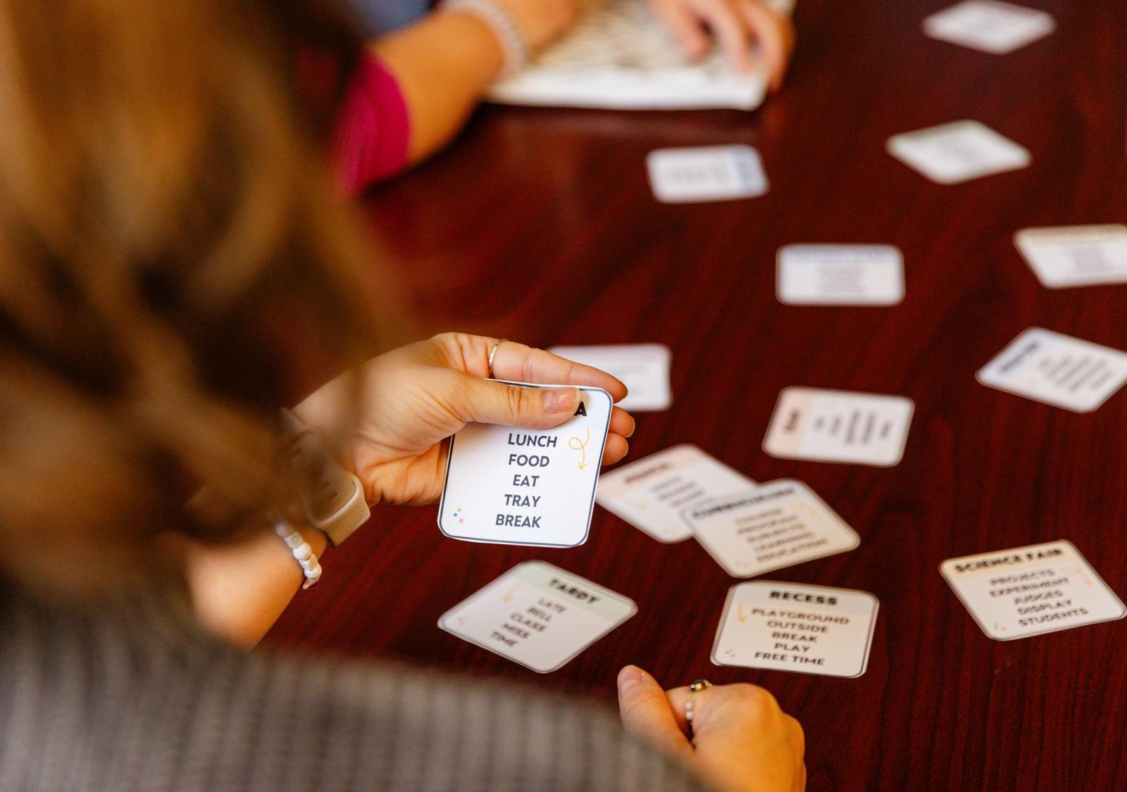 A woman sits in front of a table holding a card. Several other cards are scattered on the table in front of her.