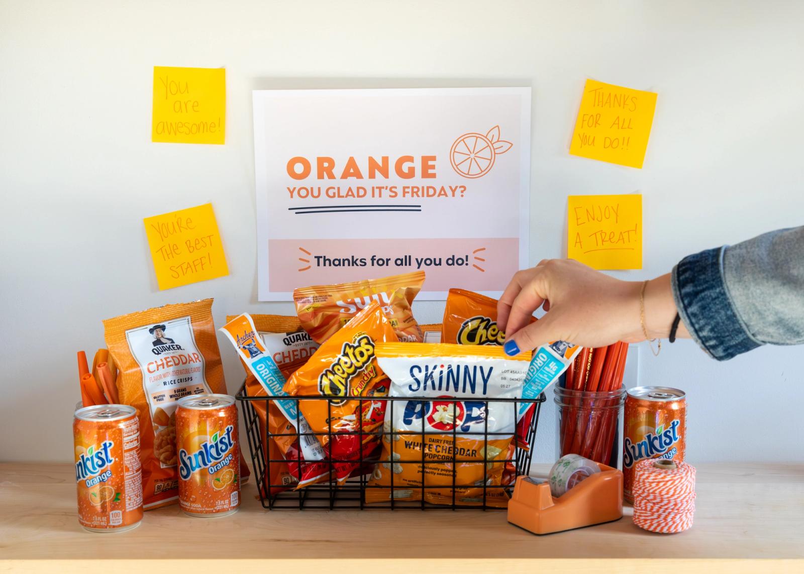 A poster that says "orange you glad it's Friday" is on a white wall with orange notes around it. Orange colored snacks are in a basket in front of it and a hand reaches in to grab one.