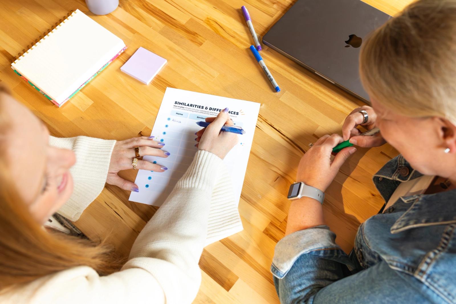 Two women sit at a wooden table. One writes on a sheet of paper. Notebooks, pens, a laptop, and post-it notes are scattered on the table.