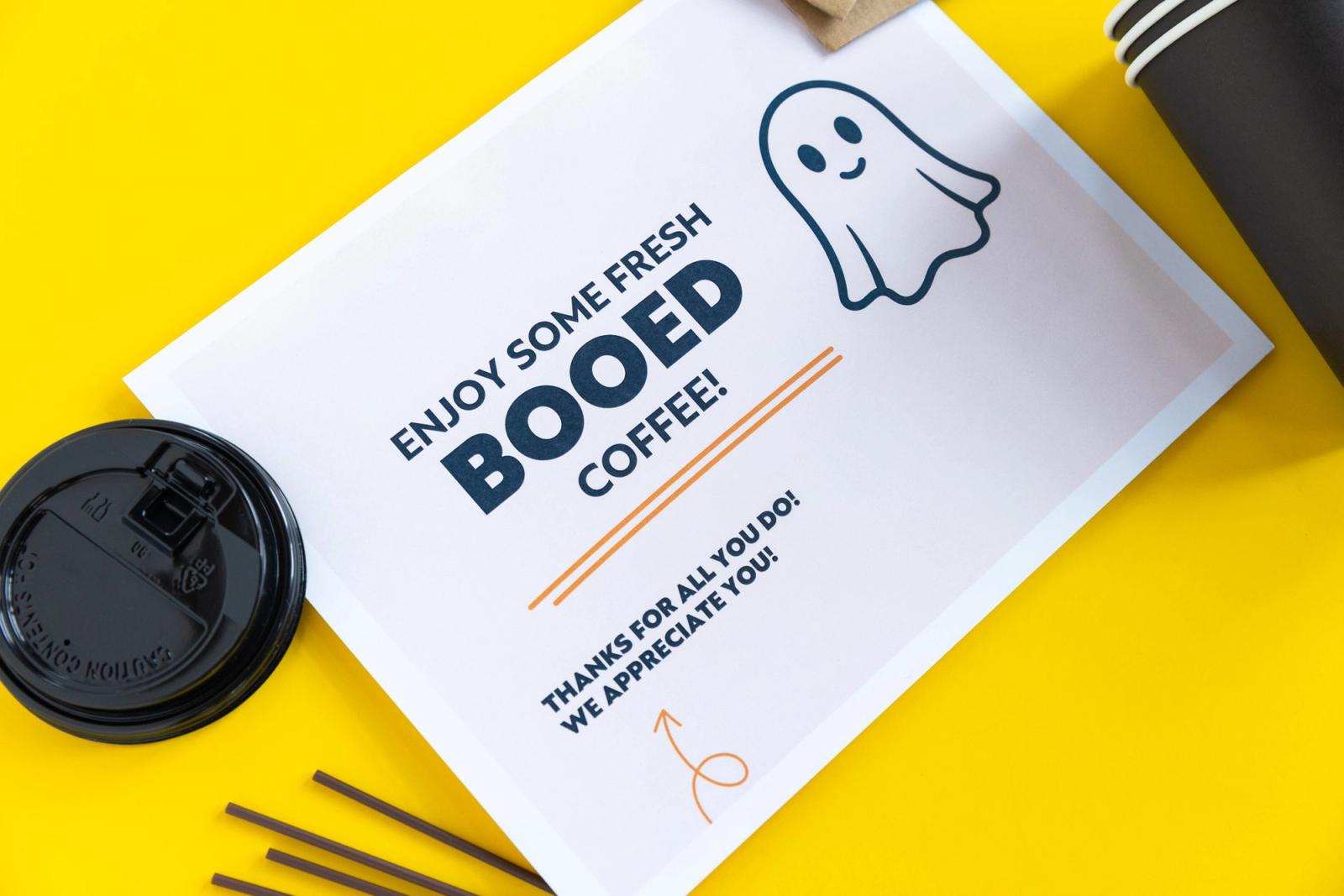 A poster that says "enjoy some freshly booed coffee" lays on a yellow background. Coffee stir sticks and to-go cup lids lay around it.