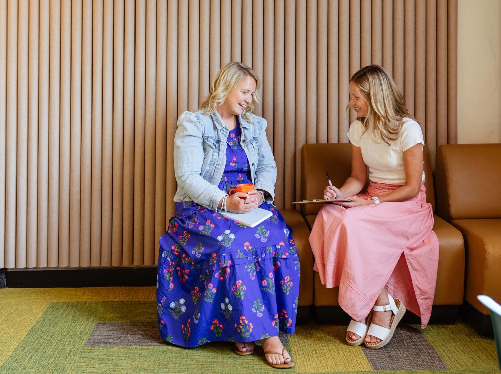 Two women sit on a couch in an office smiling and talking. One woman holds a coffee mug and one holds a clipboard and pen.