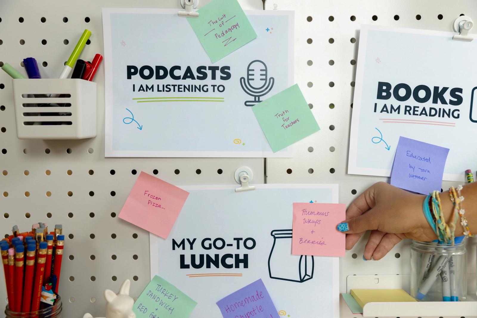Three posters with sticky notes next to them are taped to a white pegboard. A hand reaches in to place a note.