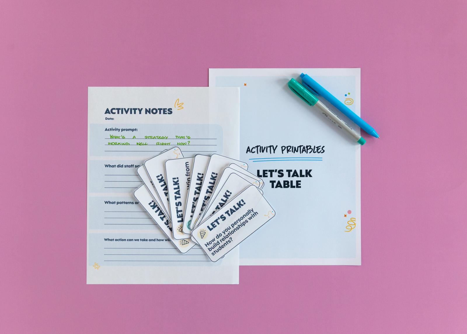 Two pieces of paper lay on a pink background. Several smaller pieces of paper fanned out and two pens lay on top. 