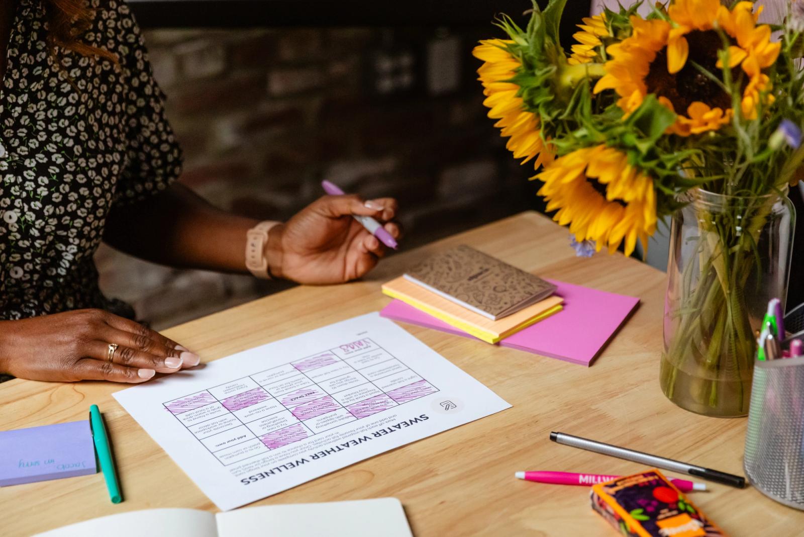 Someone sits at a desk writing on a piece of paper. Flowers, notes, and pens are on the desk.