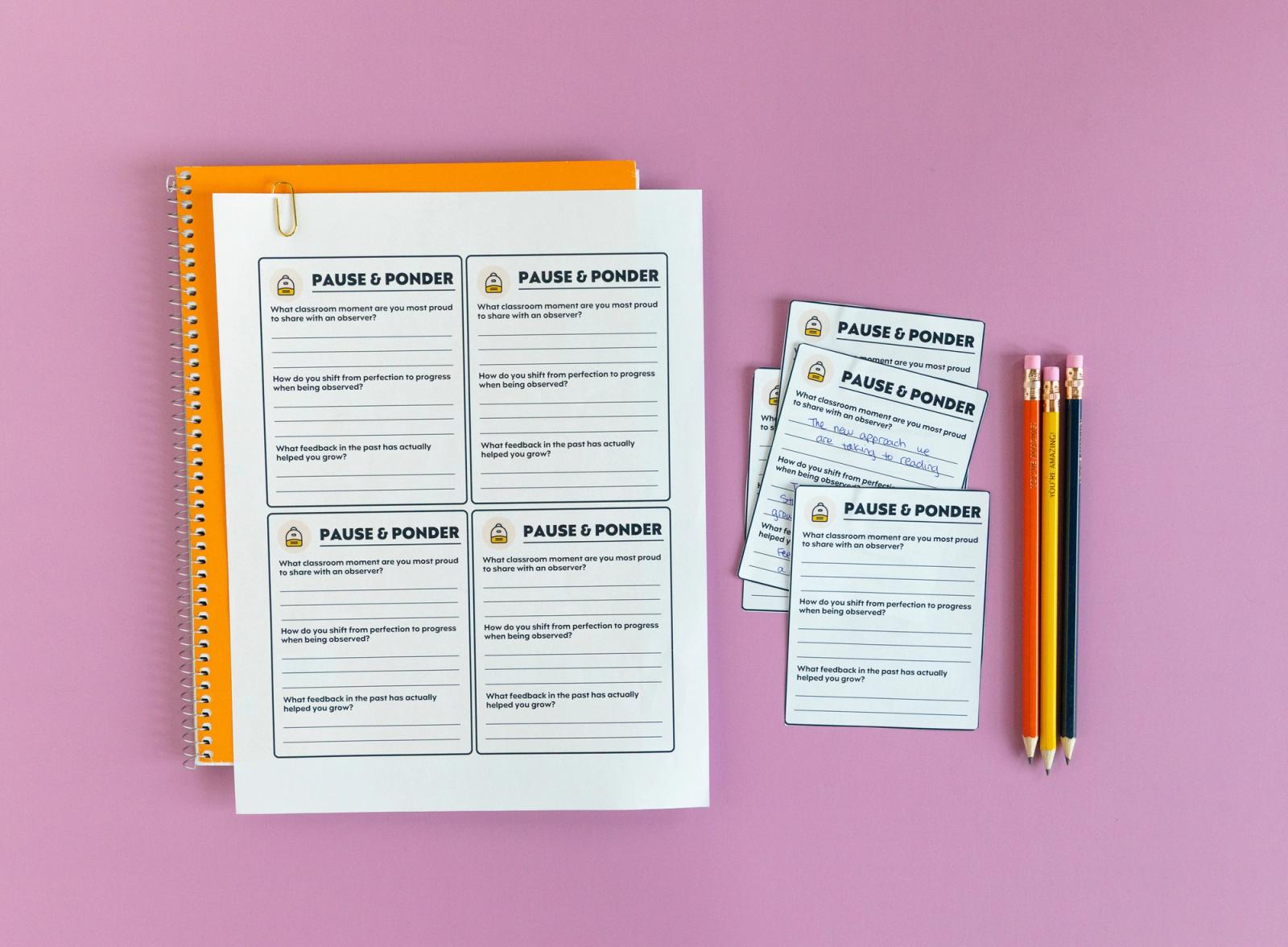 An orange notebook with a sheet of paper on it lays on a light purple background. Beside it are small notecards and three pencils.