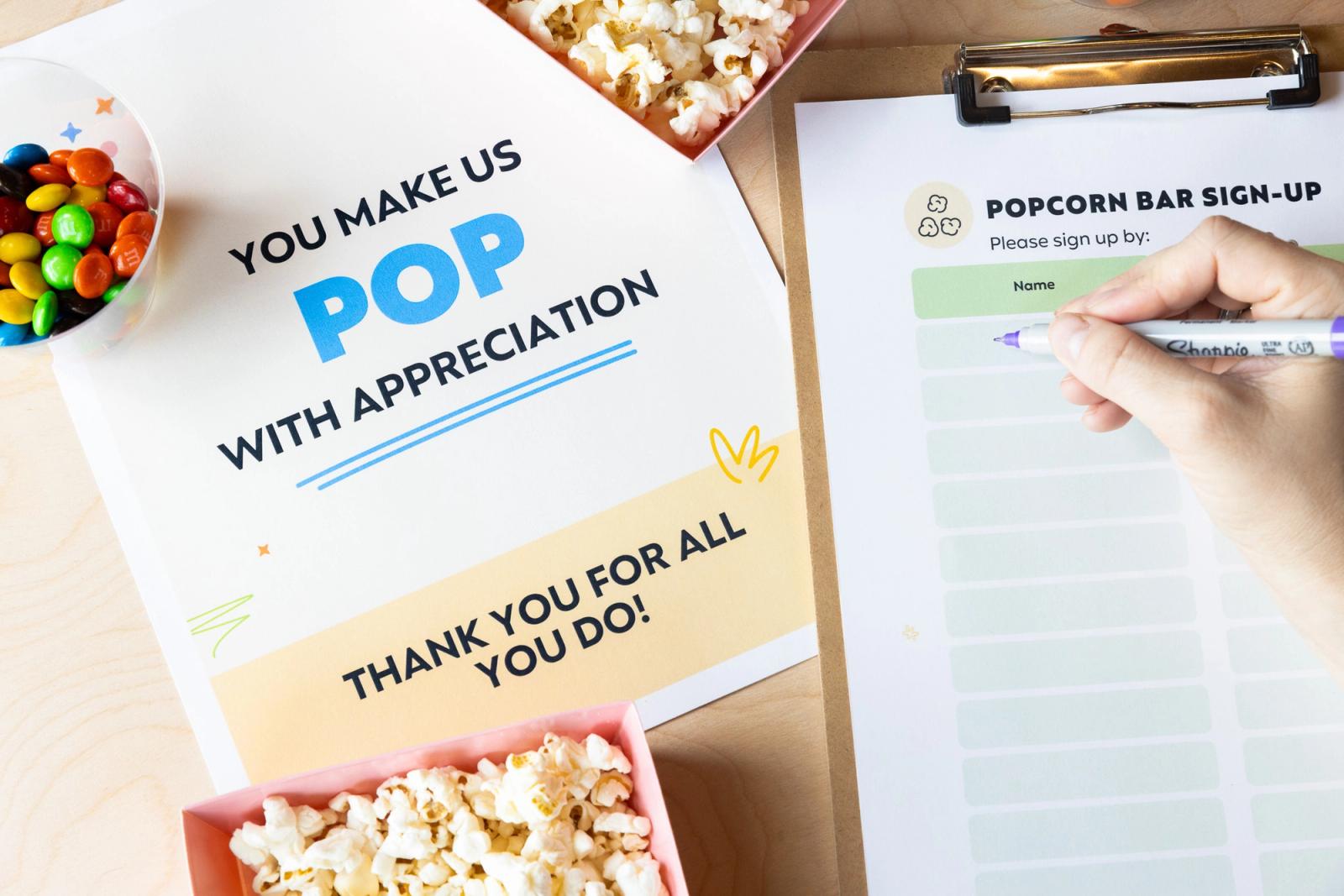 A poster, containers of popcorn and candy lay on a table. Next to it is a clipboard with a signup sheet and a hand writes on the sheet.