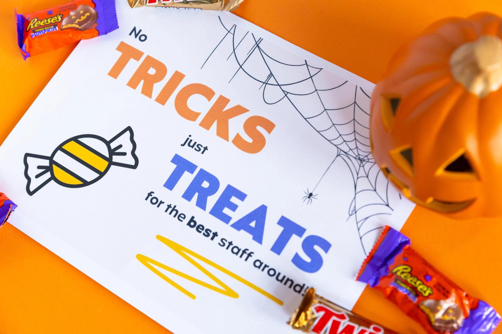 A poster that says "no tricks just treats" lays on an orange background. A pumpkin and candy lay around it.