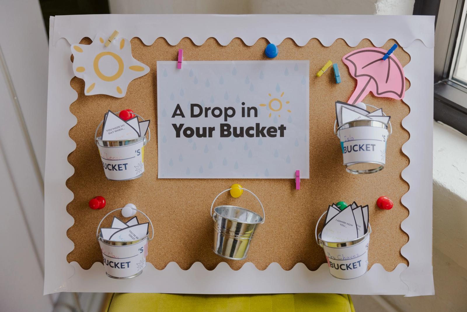 A bulletin board with a sheet of paper that says "a drop in the bucket" and small metal pails hanging on it.