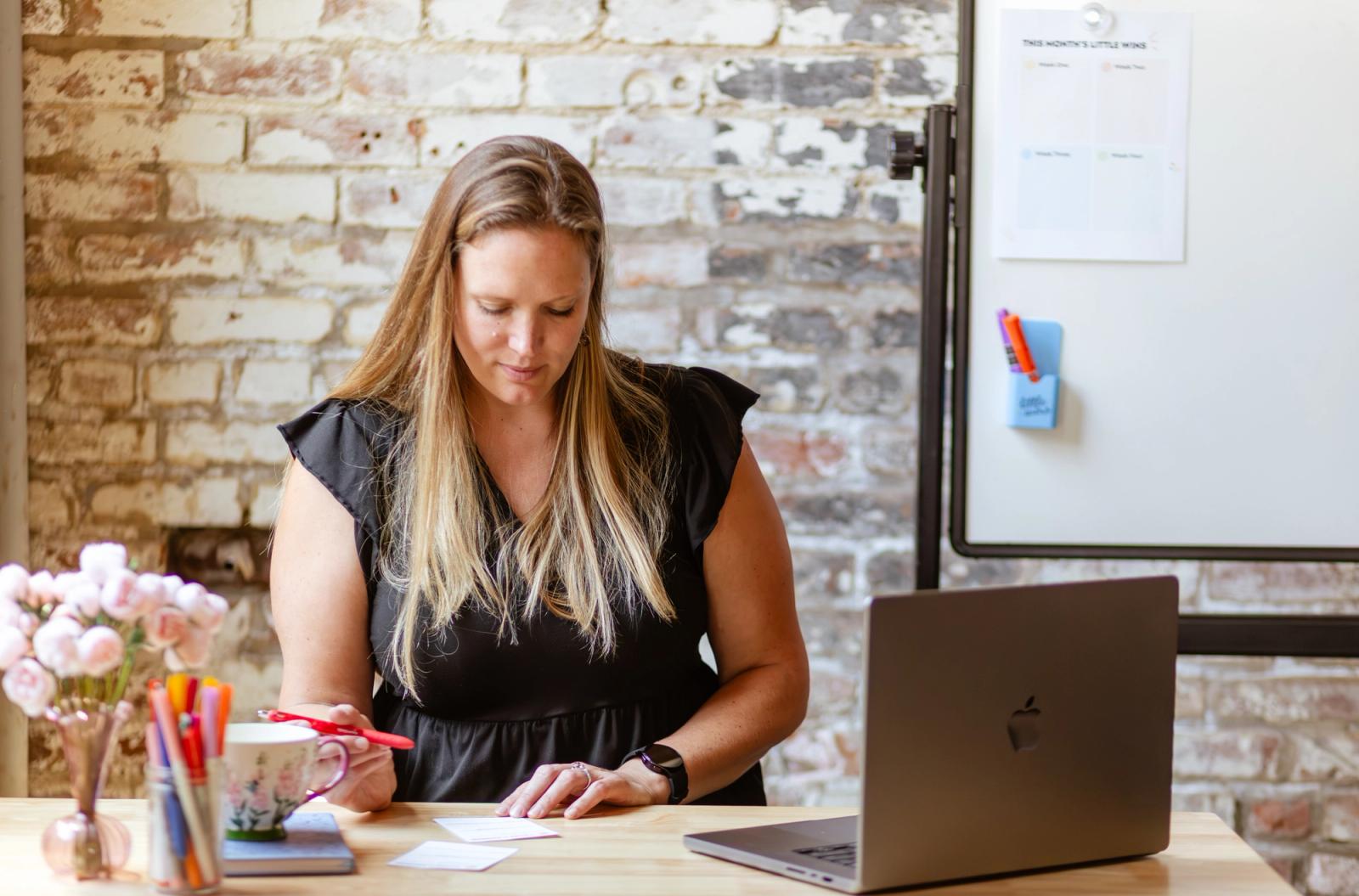 A woman sits at a desk with a laptop and vase of flows on it in front of a brick wall. She writes on notecards.