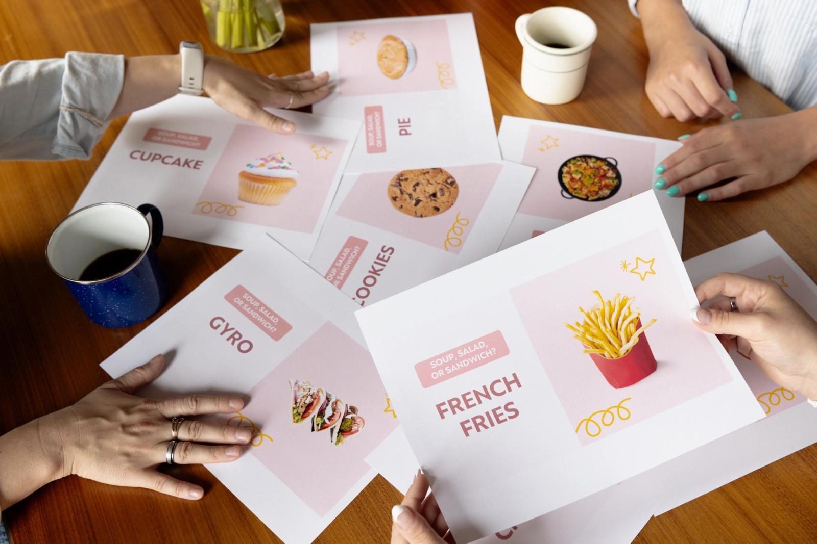 Four people sit around a table scattered with papers with images of food on them.
