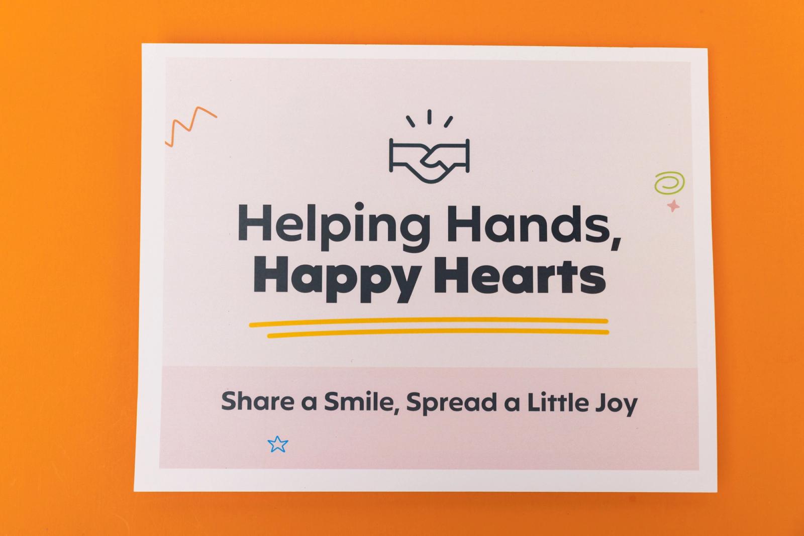 A piece of paper that says "helping hands, happy hearts" lays on an orange background.