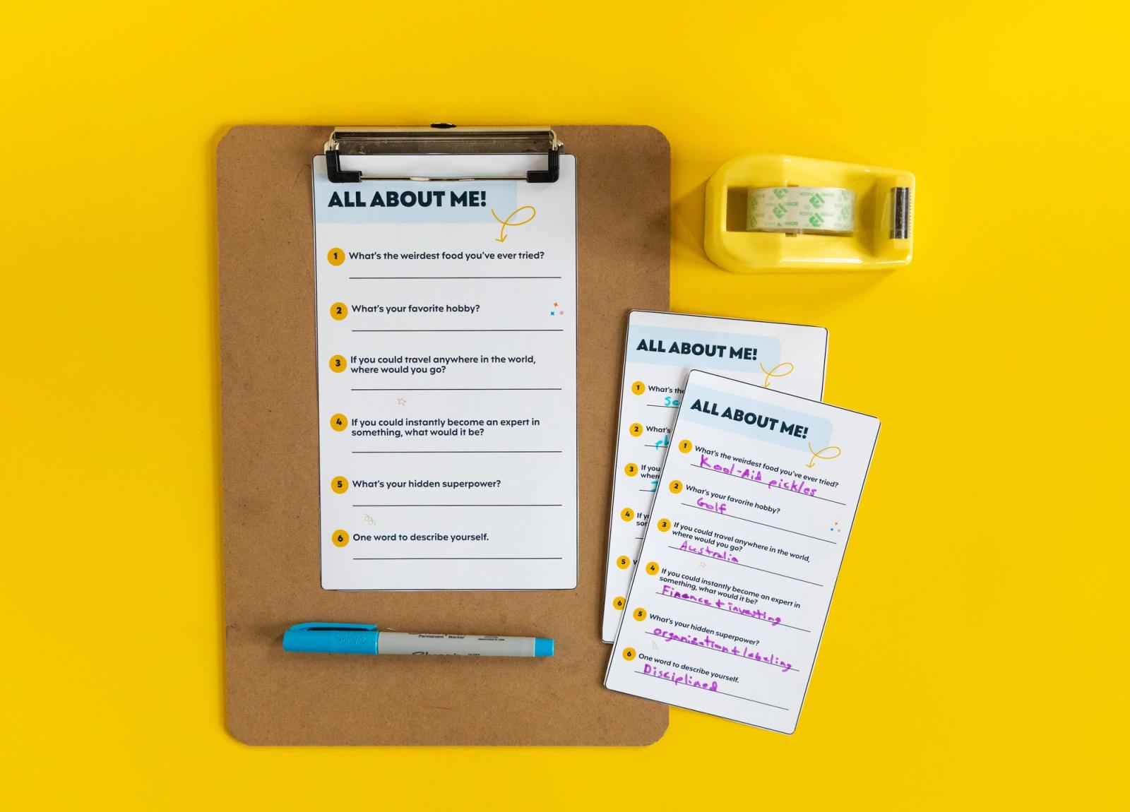 A clipboard with a small piece of paper with a list of questions sits on a yellow background. A blue pen lays on the clipboard and the right of it are more cards and a yellow tape dispenser.
