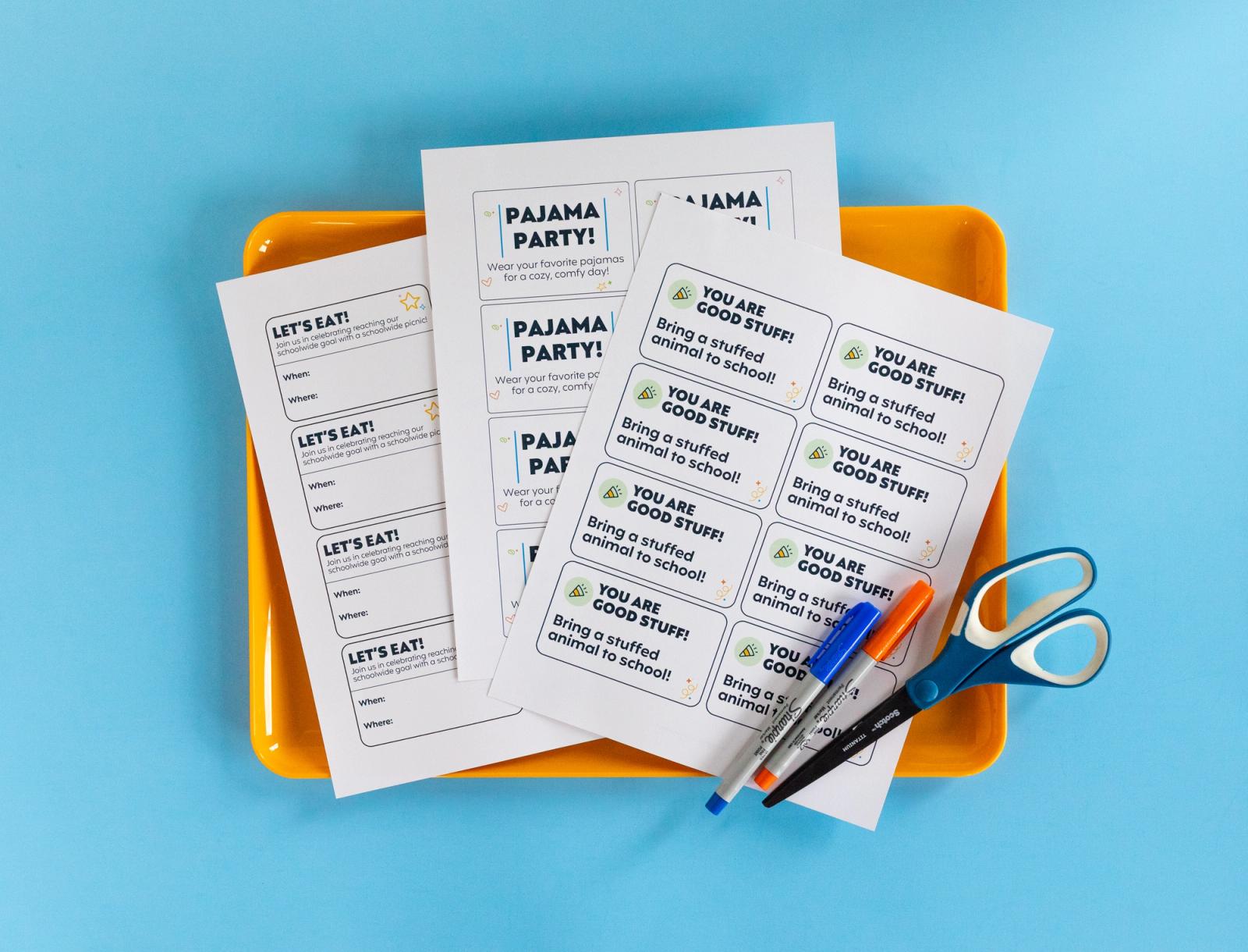 Three sheets of paper, markers, scissors lay in an orange tray on a light blue background.