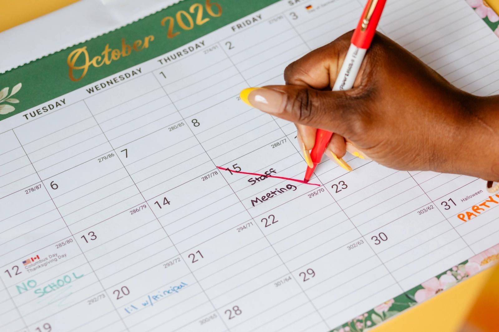A large calendar lays on a table. A hand reaches in to cross out a date that says "staff meeting."