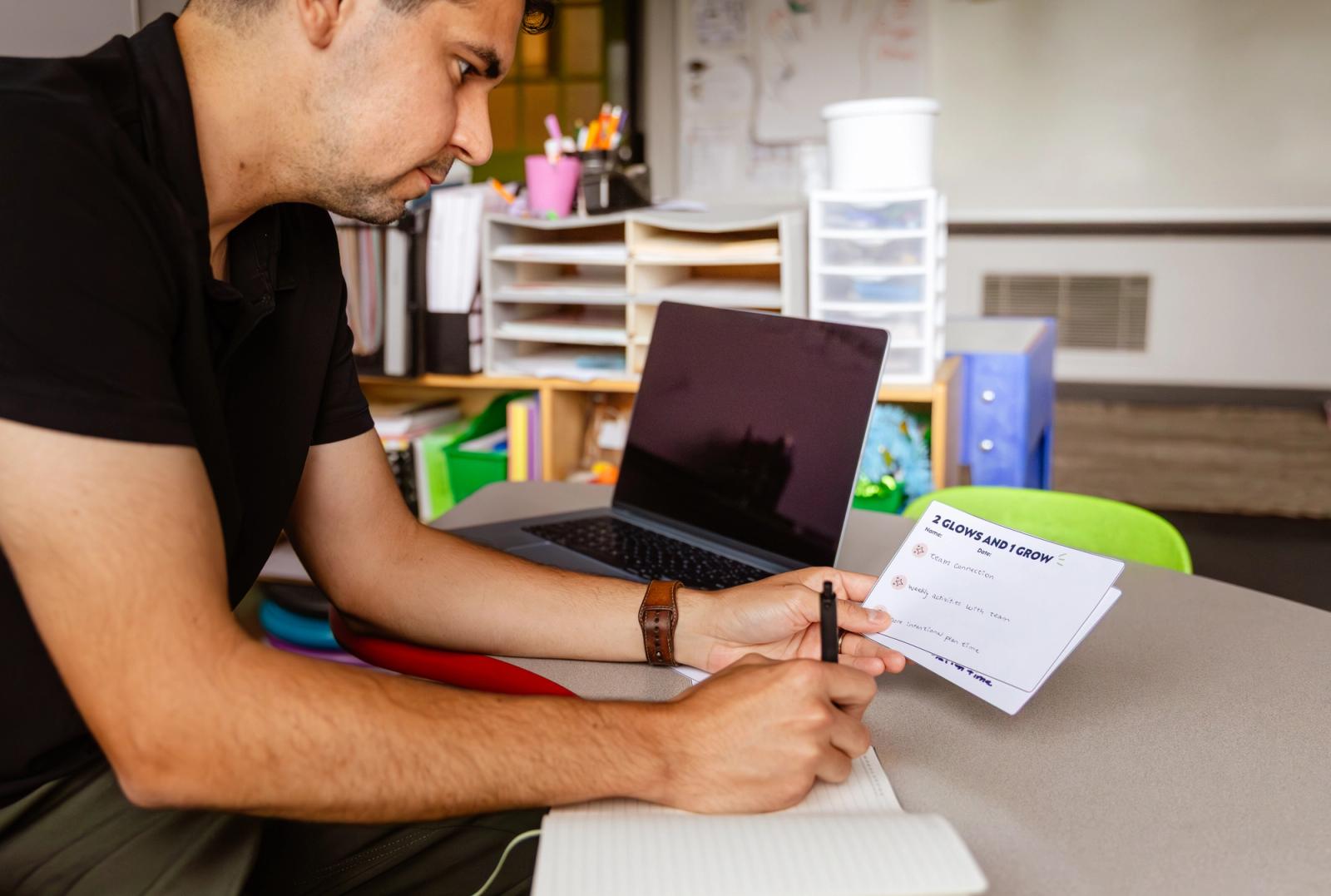 A man sits at a desk in a classroom. He is looking at blank notecards and making notes in a notebook.