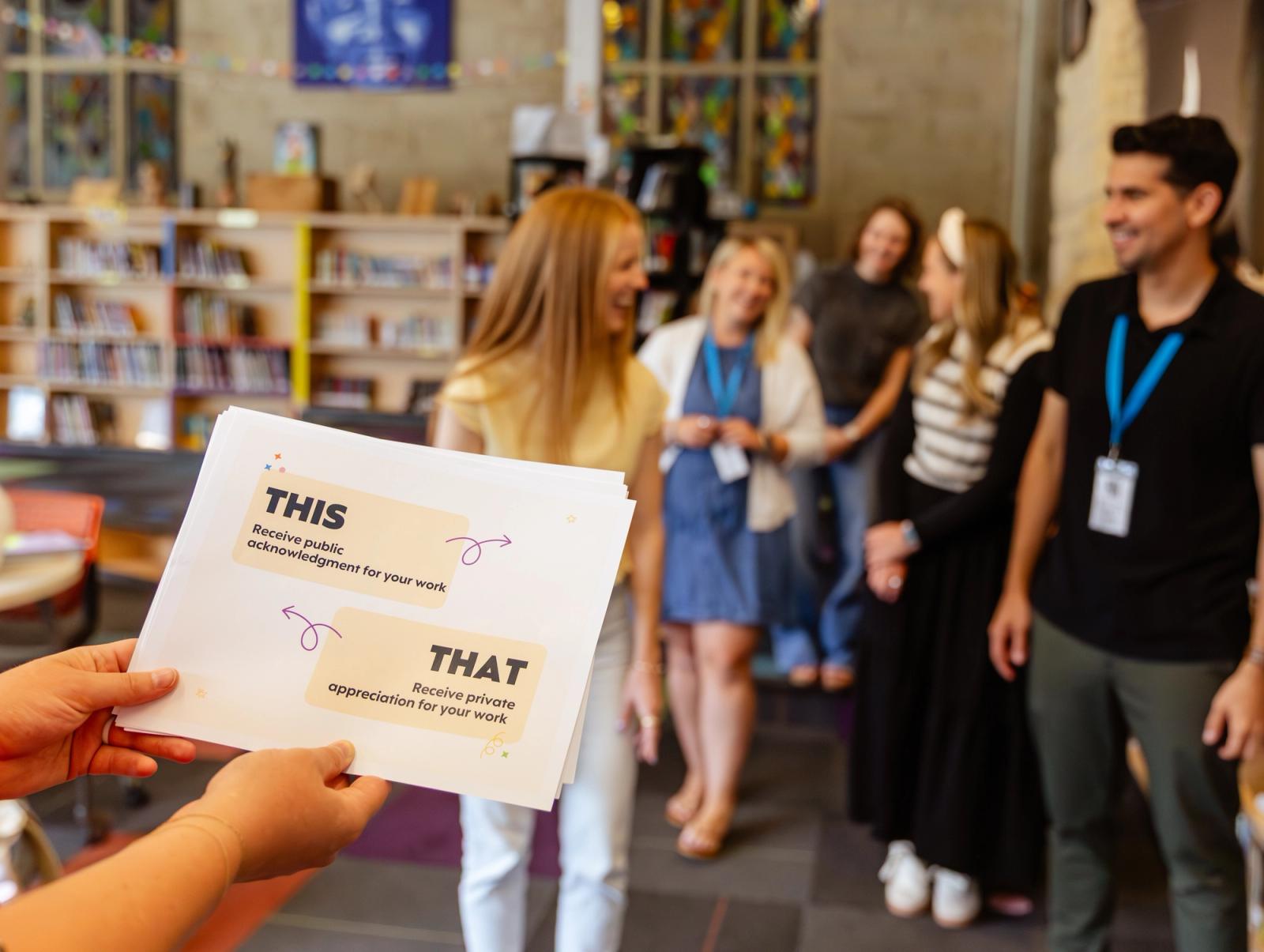 Someone holds a piece of paper with "this" and "that" on it. People in the background stand to one side of a room.