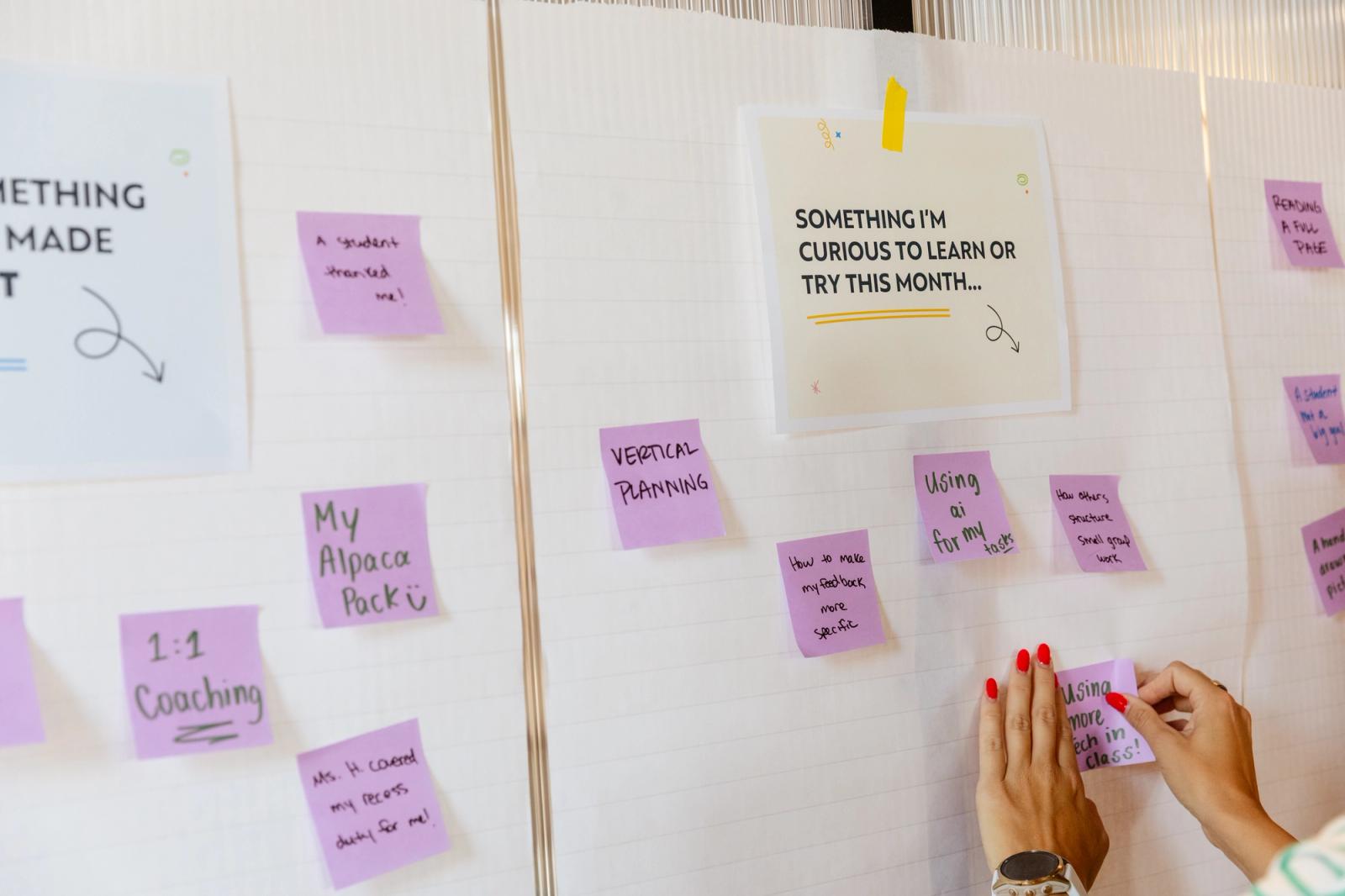 Papers are taped to a white wall with purple sticky notes stuck around them. A hand reaches in to place a note.