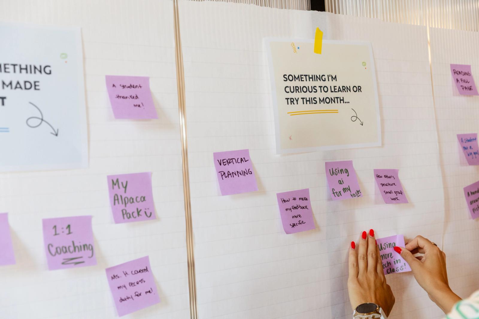 Papers are taped to a white wall with purple sticky notes stuck around them. A hand reaches in to place a note.