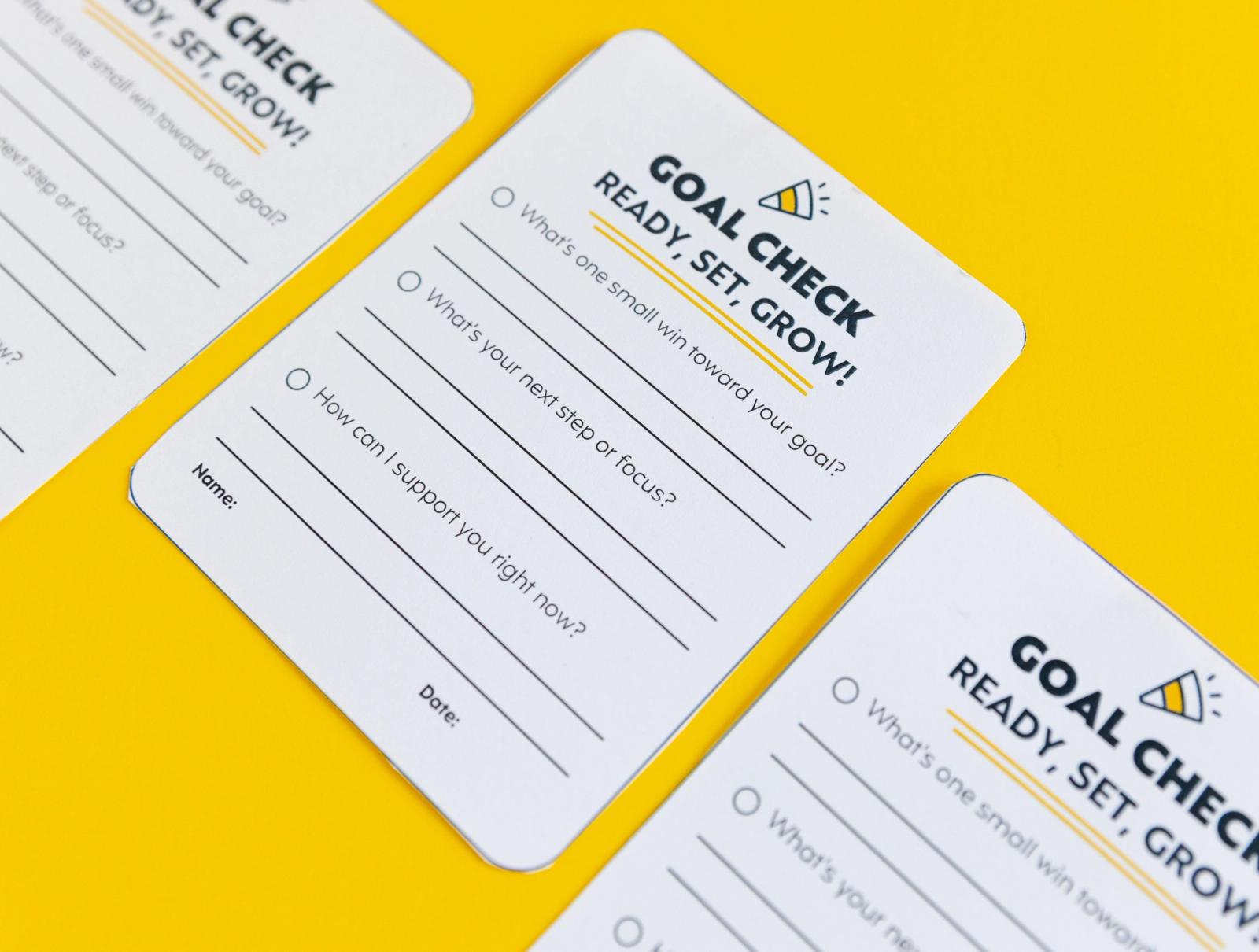 Closeup of small notecards that say "goal check" lay diagonally on a yellow background.