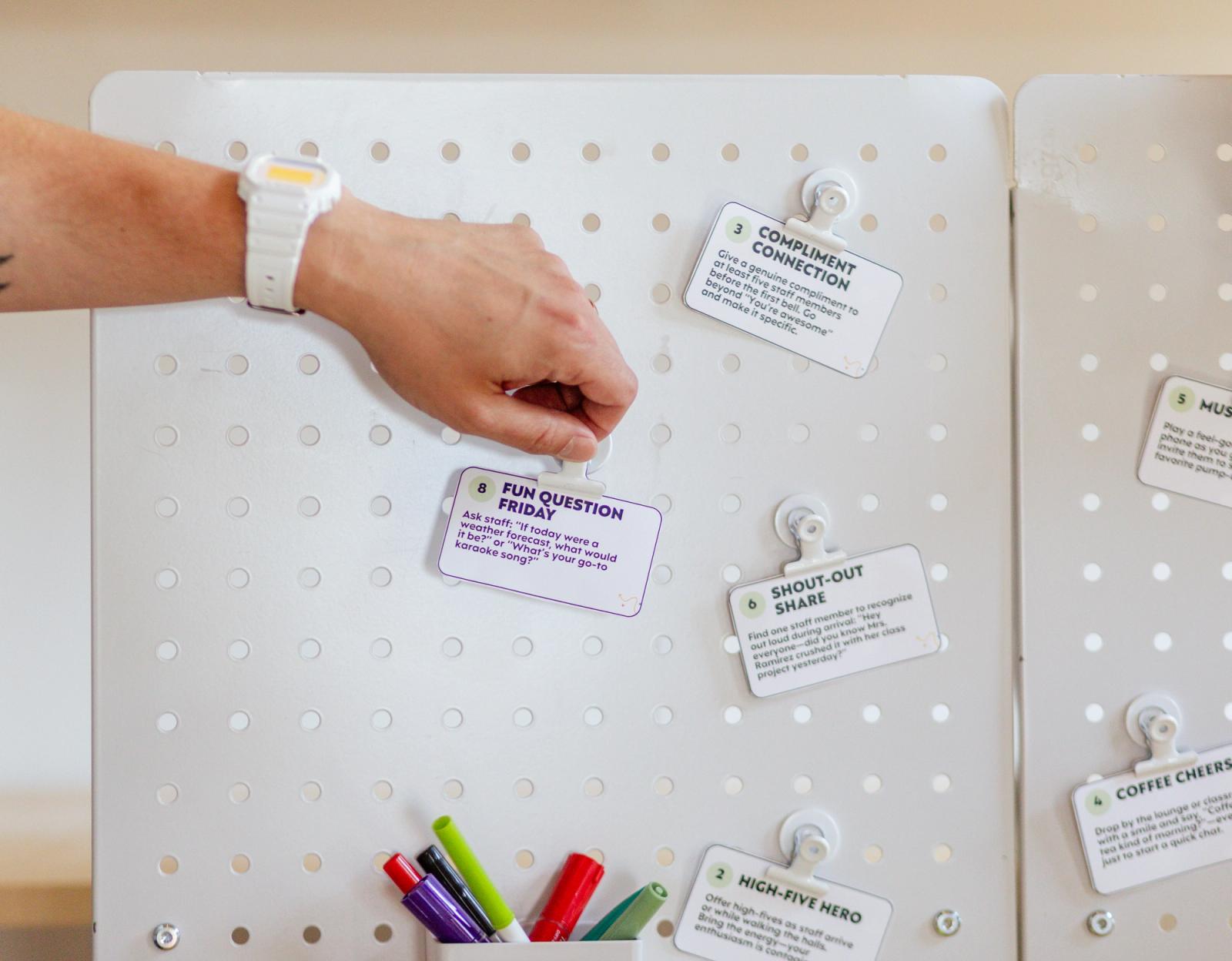 A closeup of a white peg board with small notecards on it. A hand reaches in from the left to grab a notecard.