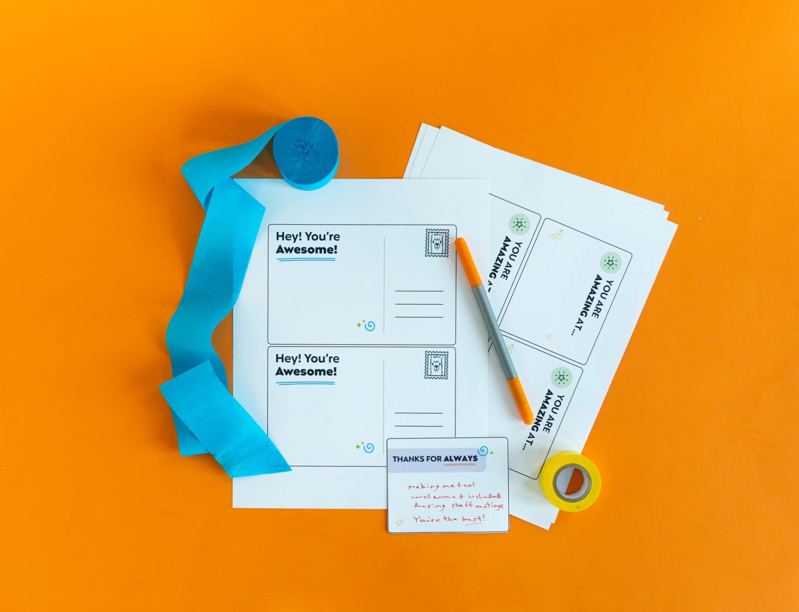 Blank postcards with positive sayings on them lay on an orange background. Beside it is a pen, toll of tape and blue streamers.