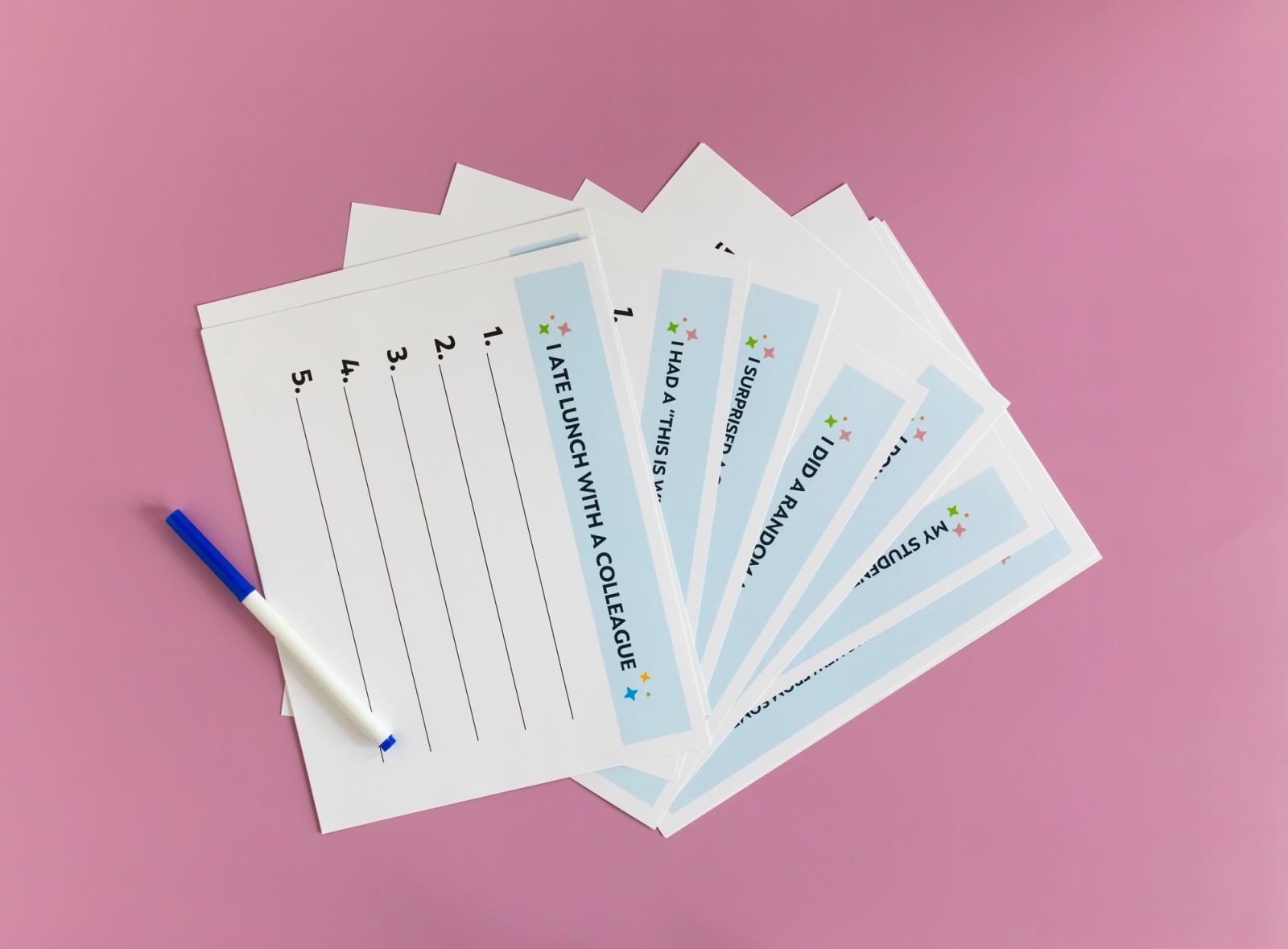 Six sheets of paper, fanned out, lay on a pink background, with a blue marker next to them.