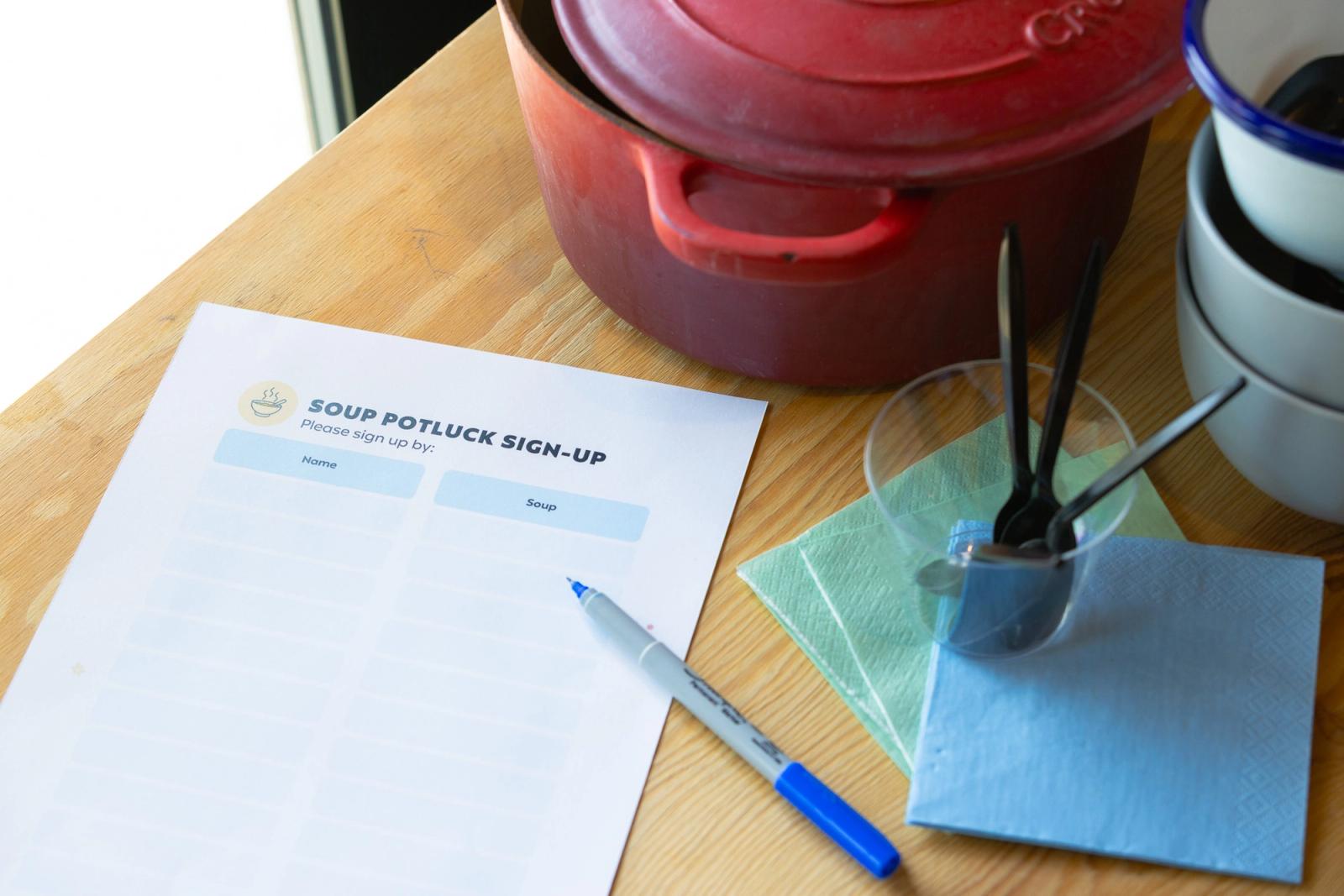 A paper sign-up sheet and pen lay on a wood table. Beside them is a red soup pan, spoons and napkins.