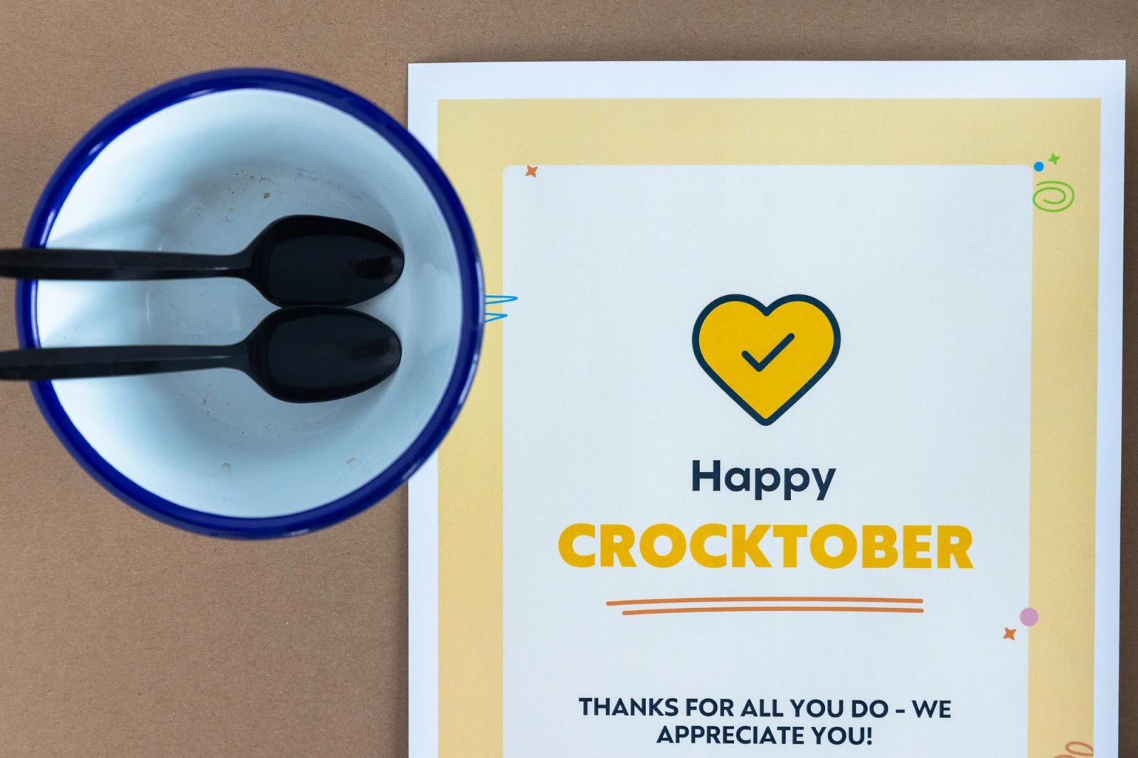 A poster with "happy crocktober" on it lays on a brown background. A bowl and spoon lays next to it.