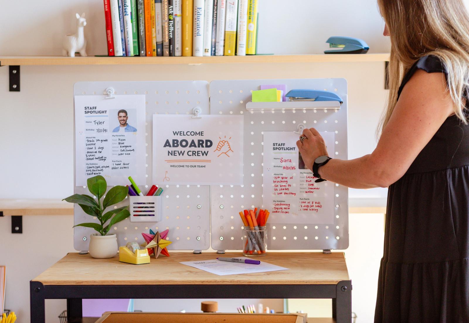 A woman stands in front of a white pegboard with posters taped to it. Assorted office supplies are on the table in front of the pegboard.