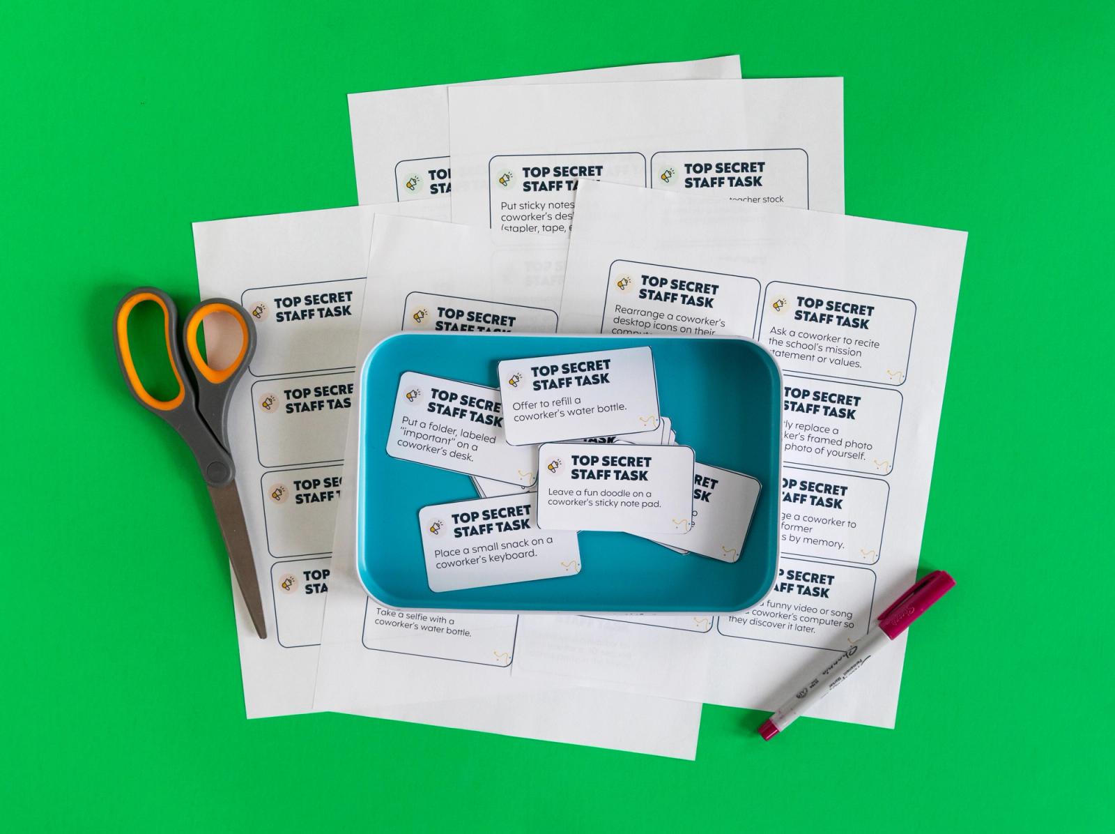 Sheets of paper, scissors, marker, and a tin with notecards in it lay on a green background.