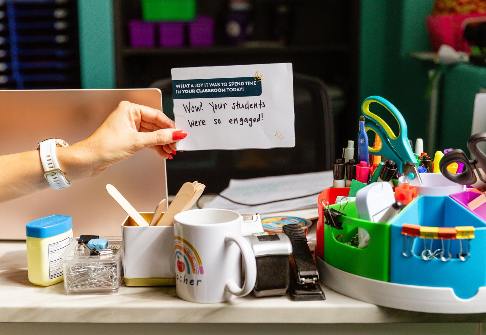 A hand reaches across a messy desk to pace a notecard near a pencil holder.