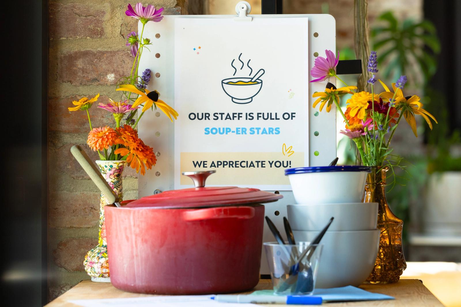 A red soup pan, bowls and spoons, and vases of flowers sit on a table in front of a poster.