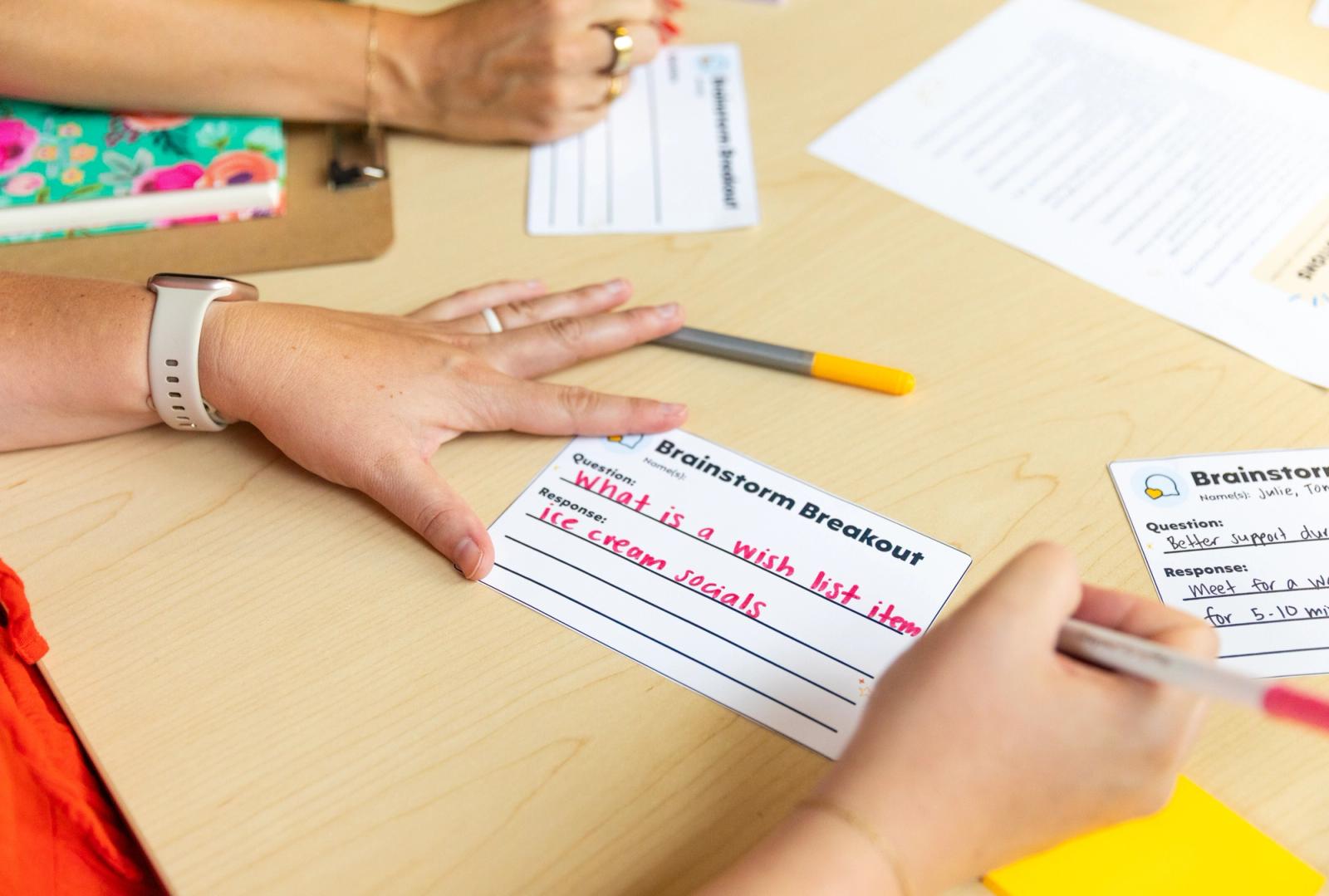 A woman writes a note on a notecard while sitting at a wooden table. Papers and pens are scattered on the table.